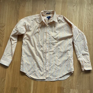 Randig skjorta från GANT, gul och vit - Säljer en snygg skjorta från GANT i gul och vit med klassiskt randigt mönster. Skjortan har lång ärm, knappar framtill och krage. Tillverkad i bomull och passar perfekt för en fräsch och stilren look.