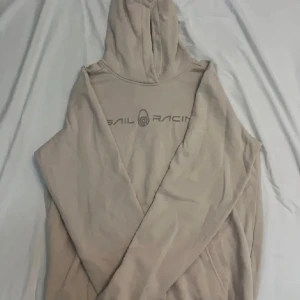Beige hoodie från Sail Racing - Beige hoodie från Sail Racing med tryckt logga på bröstet. Tröjan har huva och är långärmad, perfekt för en chill och avslappnad stil. Tillverkad i mjuk bomull som känns skön mot huden.