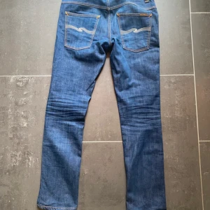Nudie Jeans blå jeans W31 L32 - Säljer ett par klassiska blå jeans från Nudie Jeans med snygga kontrastsömmar och ikoniska broderade bakfickor. Modellen har normal passform och raka ben. Tillverkade i slitstarkt bomullsjeansmaterial. Perfekta för dig som gillar stilrena och tidlösa jeans.