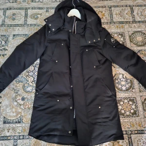 Svart parkajacka från Moose Knuckles - Svart parkajacka från Moose Knuckles (modellen heter Waterton) i nyskick med huva, flera fickor med tryckknappar och dragkedja framtill. Jackan har lång ärm och snygga detaljer på axeln. Perfekt för kalla dagar med sin stilrena look och rejäla passform.