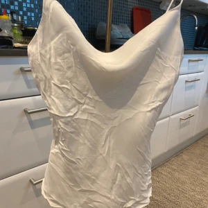 Vit satin  från Zara XS - Säljer en vit Linni från Zara i storlek XS.  har tunna axelband och en snygg draperad halsringning. Materialet är glansigt satin som ger en lyxig känsla. Perfekt för dig som gillar minimalistisk och stilren design.