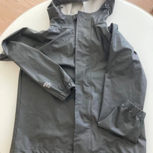 Svart regnjacka från Neomondo NYskick  - Svart regnjacka från Neomondo med huva och dragkedja framtill. Jackan har elastiska muddar vid ärmsluten och en stilren, minimalistisk look. Perfekt för blöta dagar och enkel att matcha med andra plagg.