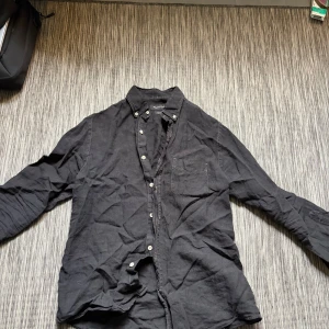 Svart linneskjorta från Massimo Dutti - Svart skjorta i 100% linne från Massimo Dutti med klassisk krage, knappar framtill och bröstficka. Skjortan har lång ärm och en stilren, tidlös look som passar perfekt till jeans eller chinos.