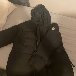 Svart pufferjacka från Tuxer XL - Svart pufferjacka från Tuxer i storlek XL med huva och dragkedja framtill. Jackan har två sidofickor med dragkedja och en snygg Tuxer-logga på ärmen. Perfekt för kalla dagar med sin vadderade design och stilrena look. Ops sönder dragkedja