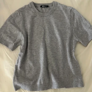 Grå stickad tröja från Gina Tricot - En enkel och stilren grå stickad tröja från Gina Tricot. Tröjan har korta ärmar, rund halsringning och ribbade kanter. Perfekt för lager-på-lager eller som den är till jeans eller kjol.