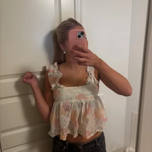 Blommig volangtopp med axelband - Supersöt blommig topp med volangdetaljer på axelbanden Ljus bas med pastelliga blommor i rosa, gul och grön.