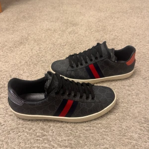 Gucci ace sneakers  - Populär svarta Gucci ace med klassiskt GG-mönster och ikoniska röd/blå ränder på sidorna. Skorna har svarta skosnören, vit sula och detaljer i rött och blått på hälen. Tillverkade i canvas och skinn för en lyxig känsla. Sicket är rätt så bra, en tvätt och de är som nya. (Kartong och påse ingår)🙌🏼🙌🏼