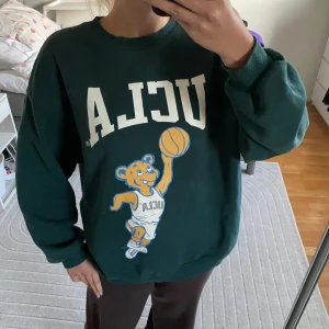 Svart UCLA sweatshirt med basketbjörn - Cool svart sweatshirt från UCLA med stort tryck av en basketspelande björn på framsidan. Tröjan har rund hals, långa ärmar och ribbade muddar. Perfekt för dig som gillar sportig och avslappnad stil.