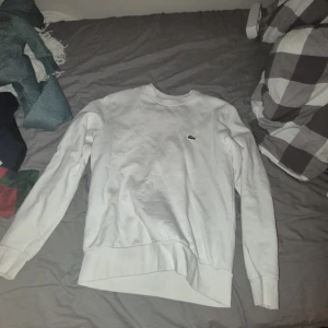 Vit sweatshirt från Lacoste - Vit sweatshirt från Lacoste med klassisk rund hals och broderad krokodil-logga på bröstet. Tröjan har lång ärm, ribbade muddar och är i en skön bomullsblandning. Perfekt för en clean och stilren look.