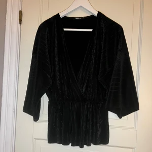 Svart omlotttopp från Gina Tricot - Snygg svart omlotttopp från Gina Tricot i mjuk sammet. Toppen har v-ringning, trekvartsärmar och markerad midja. Perfekt för dig som gillar en stilren look med lite extra lyxig känsla.