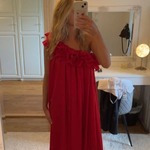 Röd oneshoulder klänning med volang - Säljer en röd långklänning med oneshoulder och stora volangdetaljer längs överdelen. Klänningen har en lös passform och faller snyggt ner mot golvet. Endast provad och prislapp sitter kvar. Perfekt för dig som vill sticka ut med en färgstark och unik look.