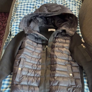 Svart pufferjacka från Moncler - Säljer en svart pufferjacka från Moncler med huva och dragkedja. Jackan har quiltad framsida i glansigt material och ärmar i ribbad textil. Fickor med tryckknappar och Moncler-logga på ärmen. Perfekt för kalla dagar.