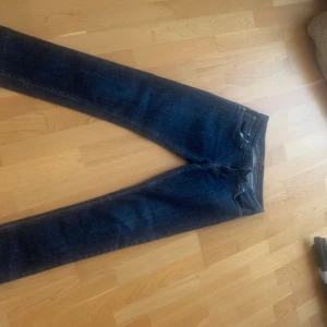 Nudie Jeans Tight Long John blå jeans - Säljer ett par mörkblå Nudie Jeans Tight Long John med klassisk orange söm och snygga bakfickor. Jeansen är i stretchigt denim, tillverkade av 97% bomull och 3% elastan. Modellen är tight och passar dig som gillar en smal siluett. Storlek 28/30 skulle jag säga, har sytt de så ignorera storleken