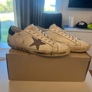 Golden Goose Superstar vita sneakers - Golden Goose Superstar sneakers köpta sommaren 2025 använda ett fåtal gånger köptes nya i göteborg för 5400kr  skor i vitt skinn med grå stjärndetalj på sidan och mörkblå mockadetalj vid hälen. Klassisk låg modell med snörning och textdetaljer på sulan. Snyggt slitna för en cool, avslappnad look. Perfekta för dig som gillar streetstyle.