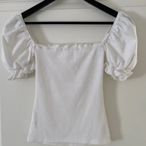 Vit puffärm topp med fyrkantig hals - Superfin vit topp med puffiga korta ärmar och fyrkantig halsringning. Toppens material har en lätt ribbad struktur och sitter snyggt på kroppen. Perfekt att styla med jeans eller kjol för en trendig look.