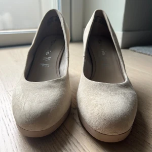 Beige mockapumps från Tamaris, strl 39 - Snygga pumps från Tamaris i beige mocka med rund tå och stadig klack. Skorna har en klassisk och stilren design med plattformsula som ger extra komfort. Perfekta för dig som vill ha en enkel men elegant look. Skada på på vänster klack, se bild 