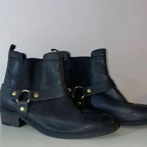 Svarta boots med gulddetaljer, stl 38 - Snygga svarta boots i skinn med guldfärgade nitar och ringar runt ankeln. Modellen har låg klack och elastiska partier på sidorna för enkel påtagning. Perfekta för dig som vill ha en cool och stilren look.