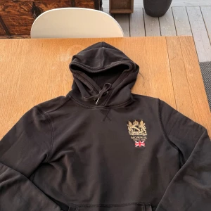 Svart hoodie från Morris med brodyr - Svart hoodie från Morris med snyggt broderat emblem och brittisk flagga på bröstet. Klassisk huva med snörning och mjukt material som känns skönt mot huden. Perfekt för dig som gillar stilrena och sportiga plagg med en touch av brittisk vibe.