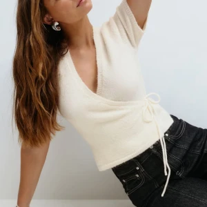 Vit omlottopp från Gina Tricot - Superfin vit omlottopp från Gina Tricot med korta ärmar och knyt i sidan. Toppen är stickad och har en v-ringad design som ger en snygg och trendig look. Perfekt att matcha med jeans eller kjol för en fräsch stil. Aldrig använd