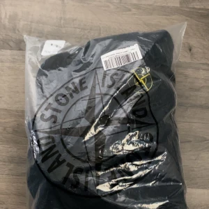 Svart zipjacka från Stone Island - Säljer en svart zipjacka från Stone Island med klassisk patch på ärmen. Jackan har huva, dragkedja framtill och två fickor. Materialet är mjukt och skönt, perfekt för dig som gillar streetwear och vill ha en clean look. Passar dig som vill ha en snygg och enkel jacka med ikonisk logga.