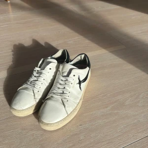 Vit-svarta sneakers från Axel Arigato - Snygga vita sneakers från Axel Arigato med svart fågelbrodyr på sidan och svart detalj vid hälen. Skorna har klassisk snörning, platt sula och är tillverkade i skinn. Perfekta för dig som gillar stilrena och trendiga sneakers.