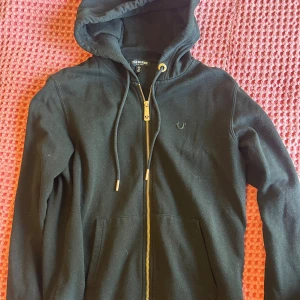 Riktigt fet truey hoodie svart - Svart hoodie från True Religion med bronzfärgad dragkedja och logga på bröstet. Tröjan har huva med snörning och känguruficka framtill. Skick 9/10. Äkta självklart