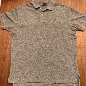 Grå pikétröja från Polo Ralph Lauren - Fet grå pikétröja från Polo Ralph Lauren med klassisk krage och två knappar framtill. Tröjan har korta ärmar och en röd broderad logga på bröstet. Perfekt för dig som gillar stilrena och tidlösa plagg med en sportig touch. Liten i storlek! 