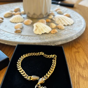 Kedjearmband i guldton - Stilrent kedjearmband i guldton med klassisk länkkedja och robust karbinlås. Armbandet har en snygg, chunky känsla och passar perfekt för dig som gillar att sticka ut med accessoarer. Levereras i svart ask.