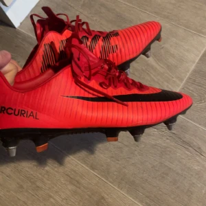 Nike Mercurial röda fotbollsskor - Säljer ett par Nike Mercurial fotbollsskor i rött med svarta detaljer och snörning. Skorna har en smal, aerodynamisk form och är designade för gräsplan med fasta dobbar. Snyggt randigt mönster och stor Nike-logga på sidan. Perfekta för dig som vill sticka ut på planen.