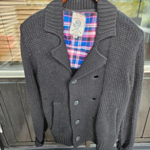 Diesel cardigan - Säljer en svart stickad kofta från Diesel med dubbelknäppning och bred krage. Koftan har långa ärmar, ribbade muddar och en snygg, klassisk look. Perfekt för kyliga dagar och passar till det mesta. Fodret har rutigt mönster i rött, blått och vitt. Hör av dig vid minsta fundering!