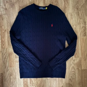 Mörkblå kabelstickad tröja Polo Ralph Lauren - Kabelstickad mörkblå tröja från Polo Ralph Lauren med klassisk rund hals och röd broderad logga på bröstet. Tröjan har långa ärmar och ribbade muddar vid ärmslut och nederkant. Perfekt för dig som gillar stilren och tidlös design. Pris kan diskuteras!