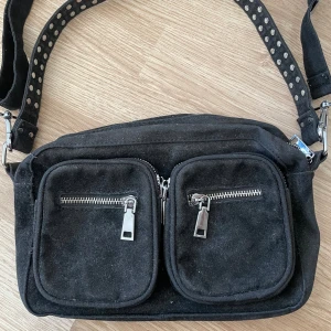 Svart axelväska med nitar från Noella - Svart axelväska från Noella i mocka med två stora framfickor och silvriga dragkedjor. De kortare axelbandet är dekorerad med silvernitar och de längre är helsvart, och även avtagbart. Perfekt storlek för att få plats med allt du behöver och flera fack inuti för bra organisering. 😚