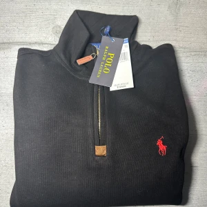 Svart half zip tröja från Polo Ralph Lauren - Svart långärmad tröja från Polo Ralph Lauren med röd broderad logga på bröstet. Tröjan har en snygg half zip-dragkedja med läderdetalj och hög krage. Tillverkad i mjuk bomull, perfekt för lager-på-lager under höst och vinter. Tröjan är ALDRIG använd! Nu pris är 125$ köpt på kryssning  