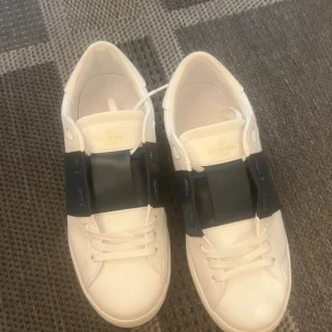 Valentino Garavani Open sneakers vit/blå - Snygga Valentino Garavani Open sneakers i vitt skinn med bred mörkblå rem över snörningen. Klassisk rund tå och platt sula med mönstrad undersida. Ikoniska nitar längs hälen ger extra edge. Perfekta för dig som gillar stilrena och lyxiga sneakers. Helt nya