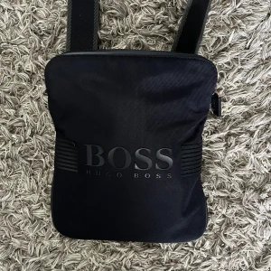 Svart axelväska från Hugo Boss - Snygg svart axelväska från Hugo Boss med justerbar rem och stilren logga på framsidan. Väskan har ett huvudfack med dragkedja och en innerficka. Materialet är slitstark polyester och designen är minimalistisk och modern.