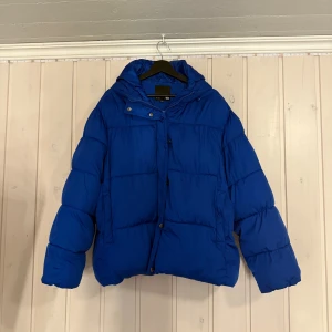 Blå pufferjacka med huva - Blå pufferjacka med huva och dragkedja samt knappar framtill. Jackan har en boxig passform och är vadderad för extra värme. Perfekt för kalla dagar och har en stilren look som passar till det mesta.