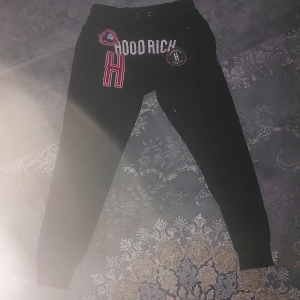 Svarta Hoodrich mjukisbyxor med patchar - Svarta mjukisbyxor från Hoodrich med vita och röda patchar och broderade detaljer framtill. Byxorna har snörning i midjan och smal passform med muddar vid bensluten. Perfekta för en streetwear-look.