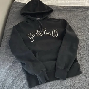 Svart hoodie från Polo Ralph Lauren - Svart hoodie från Polo Ralph Lauren med stor vit broderad POLO-text på bröstet. Klassisk huva med snörning och magficka framtill. Perfekt för en chill och stilren look. Mjuk bomullskvalitet och ribbade muddar vid ärmslut och nederkant.