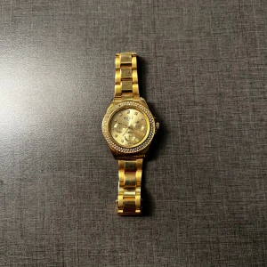 Guldig klocka från Regal med stenar - Snygg klocka från Regal i guldigt metallutförande med länkarmband. Urtavlan är guldig och dekorerad med små stenar runt boetten för extra bling. Klockan har flera små urtavlor och tydliga indexmarkeringar. Perfekt accessoar för dig som gillar att sticka ut.