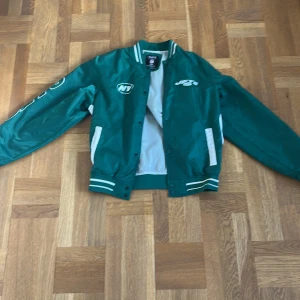 Grön NFL Jets collagejacka  - Snygg grön collagejacka från Starter med New York Jets och NFL-logga. Jackan har vita detaljer, ribbade muddar med ränder och tryckknappar framtill. Tillverkad i polyester och har klassisk sportig college-look. Perfekt för dig som vill sticka ut med amerikansk sportstil.