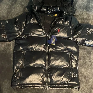 Svart pufferjacka från Polo Ralph Lauren - Snygg svart pufferjacka från Polo Ralph Lauren med glansig finish och röd broderad logga på bröstet. Jackan har huva, dragkedja framtill och två stora fickor med dragkedja. Perfekt för kalla dagar och riktigt stilren look.