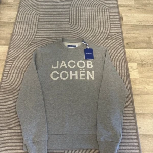 Grå sweatshirt från Jacob Cohën - Snygg grå sweatshirt från Jacob Cohën med broderad logga framtill i vitt. Tröjan har rund halsringning, långa ärmar och vanliga muddar vid ärmslut och nederkant. Perfekt för en stilren och avslappnad look. Tröjan är köpt på NK av min mamma men tyvärr var den lite för liten för mig. Den är ALDRIG använd med alla etiketter kvar på. Nypris som ni ser är 4500 men mitt pris är endast 1499 en bråkdel av priset! Skynda och fynda denna otroligt snygga och unika tröjan! Vid frågor kontakta gärna mig!