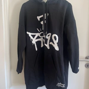 Svart Ariana Grande hoodie H&M - Svart lång  oversized hoodie från Ariana Grande x H&M med vit graffiti-inspirerad text på framsidan och tryck på ärmen. 