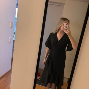 Svart omlottklänning med volang - Svart omlottklänning med v-ringning och korta, vida ärmar från Pernilla Wahlgren. Klänningen har en snygg volangkant nertill och knyts i midjan med ett band och en guldig knappdetalj. Längden är mellanlång och tyget är lätt och faller fint. Den är feltryck står att de är L men sitter som en M