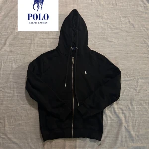 Svart hoodie från Polo Ralph Lauren - Svart hoodie från Polo Ralph Lauren med dragkedja och huva. Klassisk broderad logga på bröstet och snörning i huvan. Tillverkad i mjuk bomull med fleecefoder på insidan för extra komfort. Perfekt för en chill och stilren look.