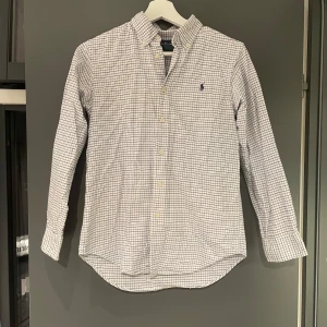 Rutig skjorta från Ralph Lauren - Klassisk vit skjorta från Ralph Lauren med blått rutmönster och broderad logga på bröstet. Skjortan har lång ärm, knappar framtill och är tillverkad i mjuk bomull. Perfekt för en clean och stilren look.