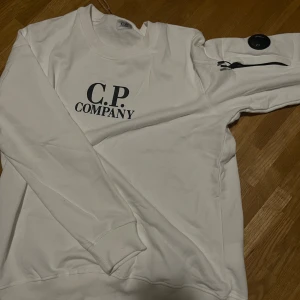 Vit sweatshirt från C.P. Company - Vit sweatshirt från C.P. Company med svart logga på bröstet och ikonisk linsdetalj på ärmen. Tröjan har rund hals, långa ärmar och ribbade muddar. Tillverkad i mjuk bomull för en clean och stilren look.