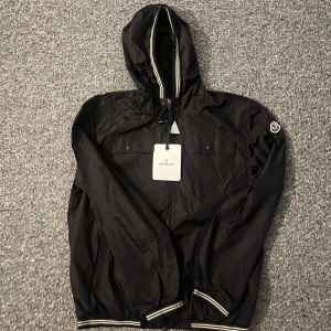 Svart vindjacka från Moncler - Svart vindjacka från Moncler med huva och dragkedja framtill. Jackan har snörning vid huvan, två bröstfickor med knappar och ribbade muddar med vita ränder. Materialet är lätt och syntetiskt, perfekt för blåsiga dagar. Moncler-logga på vänster ärm.