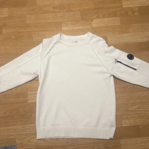 Vit stickad tröja från C.P. Company - Vit stickad tröja från C.P. Company med rund hals och ribbade muddar. På ena ärmen finns en svart detalj med logga och en snygg dragkedja. Tröjan har en clean look och passar perfekt till streetwear. Materialet är mjukt och ger en skön känsla.