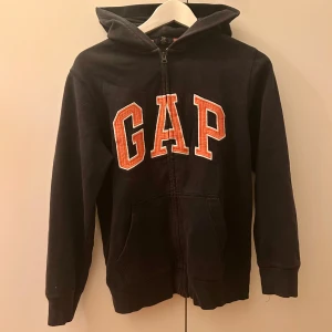 GAP hoodie - Mörkblå hoodie från GAP med stor orange logga framtill. Tröjan har huva, långa ärmar och praktisk dragkedja. Mjuk bomullskänsla och fickor på framsidan. 